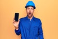 Young handsome worker man wearing uniform and hardhat holding smartphone showing screen scared and amazed with open mouth for Royalty Free Stock Photo