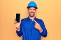 Young handsome worker man wearing uniform and hardhat holding smartphone showing screen smiling happy pointing with hand and Royalty Free Stock Photo