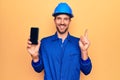 Young handsome worker man wearing uniform and hardhat holding smartphone showing screen smiling happy pointing with hand and Royalty Free Stock Photo