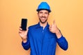 Young handsome worker man wearing uniform and hardhat holding smartphone showing screen smiling happy and positive, thumb up doing Royalty Free Stock Photo