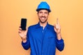 Young handsome worker man wearing uniform and hardhat holding smartphone showing screen smiling with an idea or question pointing Royalty Free Stock Photo