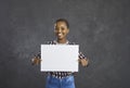 Young happy african american woman holding a blank white sheet while standing on a gray background. Royalty Free Stock Photo