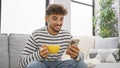 Young, happy arab man relaxing at home, enjoying his coffee while texting on his smartphone Royalty Free Stock Photo