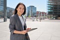 Young happy Asian business woman standing on city street holding digital tablet.