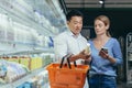 Young happy Asian couple using smartphone in supermarket with shopping cart choosing products while grocery store Royalty Free Stock Photo