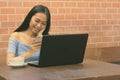 Young beautiful Asian teenage girl sitting while using laptop against brick wall Royalty Free Stock Photo