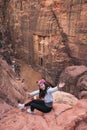 Young happy Asian woman traveller with smiley face sitting on rock at viewpoint of Petra ruin and ancient city looking at the