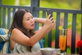 Young happy and attractive Asian Chinese woman at outdoors cafe drinking juice taking selfie self portrait with mobile phone at Royalty Free Stock Photo