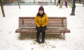 Young happy and attractive Asian Korean woman in Winter jacket and beanie enjoying snowfall at city park sitting on bench