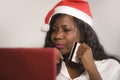 Young happy and attractive black African American woman in Santa Claus hat holding credit card buying Christmas present and gift o Royalty Free Stock Photo