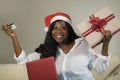 Young happy and attractive black African American woman in Santa Klaus hat holding credit card and Christmas present box doing onl Royalty Free Stock Photo