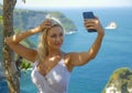 Young happy and attractive blond woman taking selfie portrait with mobile phone at beautiful tropical paradise view of ocean rock Royalty Free Stock Photo