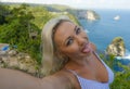 Young happy and attractive blond woman taking selfie portrait with mobile phone at beautiful tropical paradise view of ocean rock Royalty Free Stock Photo