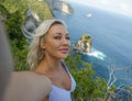 Young happy and attractive blond woman taking selfie portrait with mobile phone at beautiful tropical paradise view of ocean rock Royalty Free Stock Photo