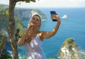 Young happy and attractive blond woman taking selfie portrait with mobile phone at beautiful tropical paradise view of ocean rock Royalty Free Stock Photo