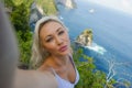 Young happy and attractive blond woman taking selfie portrait with mobile phone at beautiful tropical paradise view of ocean rock Royalty Free Stock Photo