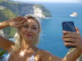 Young happy and attractive blond woman taking selfie portrait with mobile phone at beautiful tropical paradise view of ocean rock Royalty Free Stock Photo