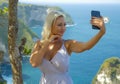 Young happy and attractive blond woman taking selfie portrait with mobile phone at beautiful tropical paradise view of ocean rock Royalty Free Stock Photo
