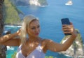 Young happy and attractive blond woman taking selfie portrait with mobile phone at beautiful tropical paradise view of ocean rock Royalty Free Stock Photo