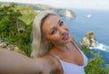 Young happy and attractive blond woman taking selfie portrait with mobile phone at beautiful tropical paradise view of ocean rock Royalty Free Stock Photo