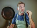 Young happy and attractive home cook man holding cooking pan smiling satisfied and proud giving delicious hand fingers sign as dom