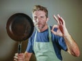 Young happy and attractive home cook man holding cooking pan smiling satisfied and proud giving okay sign on isolated background