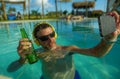 Young happy and attractive man taking selfie picture with mobile phone drinking beer and listening to music at tropical resort Royalty Free Stock Photo