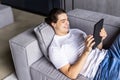 Attractive man using digital pad or tablet lying relaxed on couch at home connected to internet reading , watching the screen and Royalty Free Stock Photo