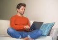 Young happy and attractive man working relaxed with laptop computer at modern apartment living room sitting at sofa couch typing a Royalty Free Stock Photo