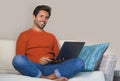 Young happy and attractive man working relaxed with laptop computer at modern apartment living room sitting at sofa couch typing a Royalty Free Stock Photo