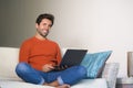 Young happy and attractive man working relaxed with laptop computer at modern apartment living room sitting at sofa couch typing a