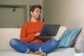 Young happy and attractive man working relaxed with laptop computer at modern apartment living room sitting at sofa couch typing a Royalty Free Stock Photo