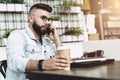 Young happy bearded man having mobile phone conversation with business partner, while sitting with laptop in cafe. Royalty Free Stock Photo