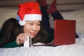 Young happy and beautiful Asian American girl on bed in Santa Christmas hat using credit card and laptop computer for online shopp Royalty Free Stock Photo