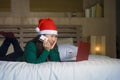 Young happy and beautiful Asian Chinese girl on bed in Santa Christmas hat using credit card and laptop computer for online shoppi Royalty Free Stock Photo