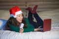 Young happy and beautiful Asian Chinese girl on bed in Santa Christmas hat using credit card and laptop computer for online shoppi Royalty Free Stock Photo