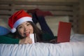 Young happy and beautiful Asian Chinese girl on bed in Santa Christmas hat using credit card and laptop computer for online shoppi Royalty Free Stock Photo