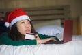 Young happy and beautiful Asian Chinese woman on bed in Santa Christmas hat using credit card and laptop computer for online shopp Royalty Free Stock Photo