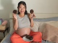Young happy and beautiful Asian Chinese woman pregnant sitting on bed holding dish full of chocolate donuts craving for sweet food Royalty Free Stock Photo