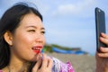 Young happy and beautiful Asian Chinese woman retouching her makeup applying red lipstick on her lips using mobile phone as mirror Royalty Free Stock Photo