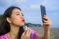 Young happy and beautiful Asian Chinese woman retouching her makeup applying red lipstick on her lips using mobile phone as mirror Royalty Free Stock Photo