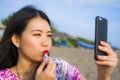 Young happy and beautiful Asian Chinese woman retouching her makeup applying red lipstick on her lips using mobile phone as mirror Royalty Free Stock Photo