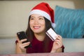 Young happy and beautiful Asian Chinese woman in Santa Claus hat holding credit card using mobile phone for Christmas online shopp Royalty Free Stock Photo