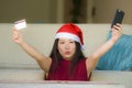 Young happy and beautiful Asian Chinese woman in Santa Claus hat holding credit card using mobile phone for Christmas online shopp Royalty Free Stock Photo