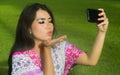 Young happy and beautiful Asian Chinese woman taking selfie pic with mobile phone camera throwing a kiss posing outdoors on green