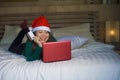 Young happy and beautiful Asian Korean girl on bed in Santa Christmas hat using credit card and laptop computer for online shoppin Royalty Free Stock Photo