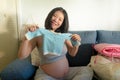 Young happy and beautiful Asian Korean woman pregnant on couch enjoying playful with little clothes of upcoming baby smiling