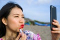 Young happy and beautiful Asian Korean woman retouching her makeup applying red lipstick on her lips using mobile phone as mirror Royalty Free Stock Photo