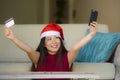 Young happy and beautiful Asian Korean woman in Santa Claus hat holding credit card using mobile phone for Christmas online shoppi Royalty Free Stock Photo