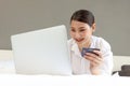 Young happy beautiful Asian woman using laptop computer and holding credit card while lying on the bed, businesswoman working Royalty Free Stock Photo